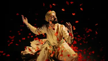 A man in traditional Persian attire gracefully dances amidst swirling red rose petals against a dark background.