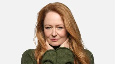 A woman with long red hair wearing a green sweater, holding her hands up under her face against a white background.