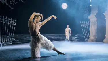 A topless man in a white feathery costume dancing under a set with a full moon on a blue lit stage.