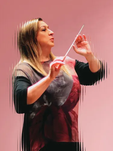 Simone Young standing side on, holding a conductor's baton, against a pink background.
