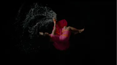A birdseye view of a white woman in a pink and orange dress laying in shallow water on a black ground.