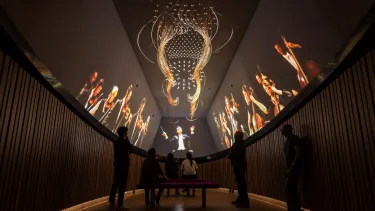 A group of visitors watching a projection of an orchestra show on ceiling screens.