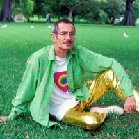 A white man in his 50s wearing gold pants and a green and red spotty shirt sits relaxed on grass.