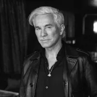 Black and white portrait of a man with white hair and a mustache, wearing a leather jacket and pendant. He exudes confidence, with a dimly lit background.
