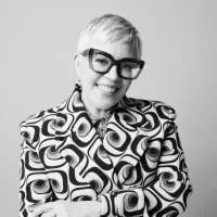 Black and white image of a smiling person with short hair, wearing large glasses and a bold, patterned jacket with swirling designs. Confident and cheerful.