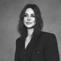 Black and white portrait of a woman with wavy hair, wearing a dark suit jacket and a subtle smile, conveying confidence and poise against a gray background.