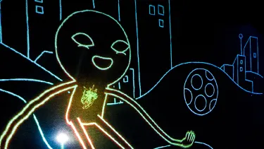 A woman in striped shirt and overalls stands on stage with a glowing stick figure drawing.